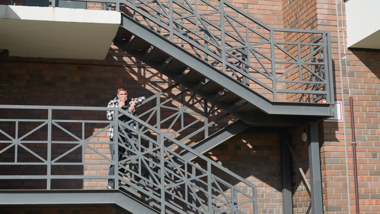 Student in black and white shirt smokes cigarette while leaning on metal walkway railing of brick building under bright sunlight with geometric shadows and urban structure in background