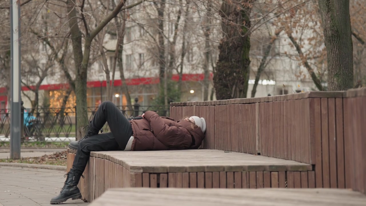 Mulher caminhando e depois reclinando em banco de madeira em parque da cidade, postura relaxada com as pernas elevadas, casaco acolchoado e gorro de tricô, árvores sem folhas e fundo urbano suave, humor calmo e introspectivo
