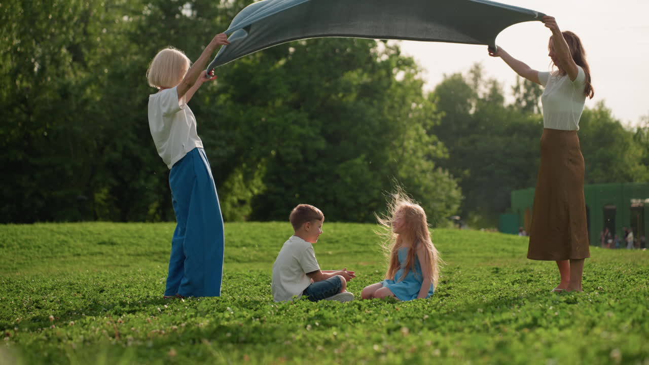 Toddlers seated on grass while parents spread blanket over them in sunny park, face painted girl and boy sharing laughter, family bonding during playful summer picnic on green clover field