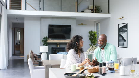 Feliz-Pareja-Birracial-Desayunando-Usando-Un-Teléfono-Inteligente-Y-Hablando-En-La-Cocina,-Cámara-Lenta