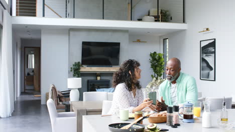 happy biracial couple having breakfast using smartphone and talking in kitchen, slow motion