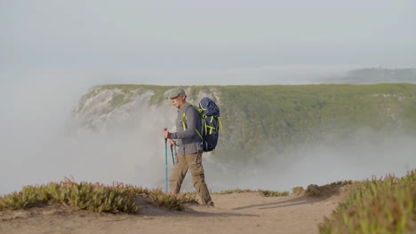 long shot of focused man with backpack climbing mountain with trekking poles alone on a foggy day