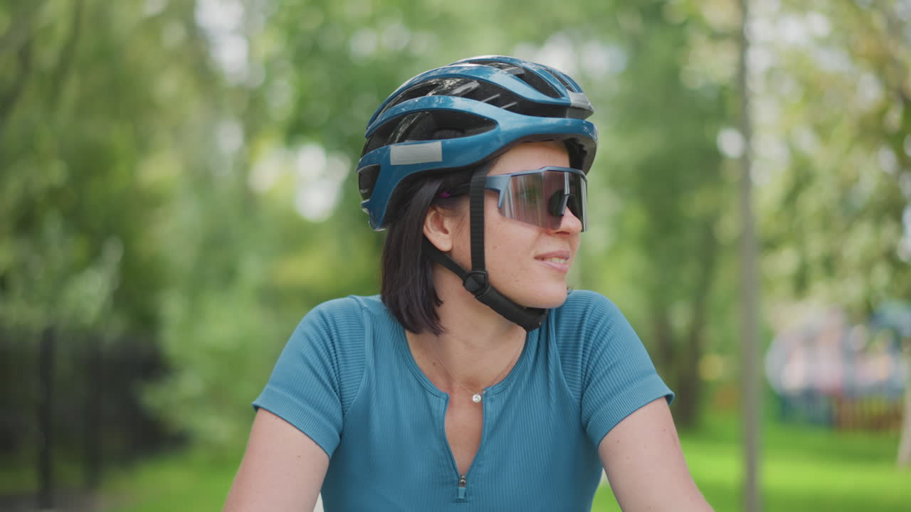 Ciclista usando capacete e óculos de sol, sorrindo enquanto faz uma pausa na trilha do parque, close-up com viseira reflexiva, camiseta verde-água, cabelo curto, estilo casual de quem usa para se locomover, aproveitando a luz do sol, contato visual