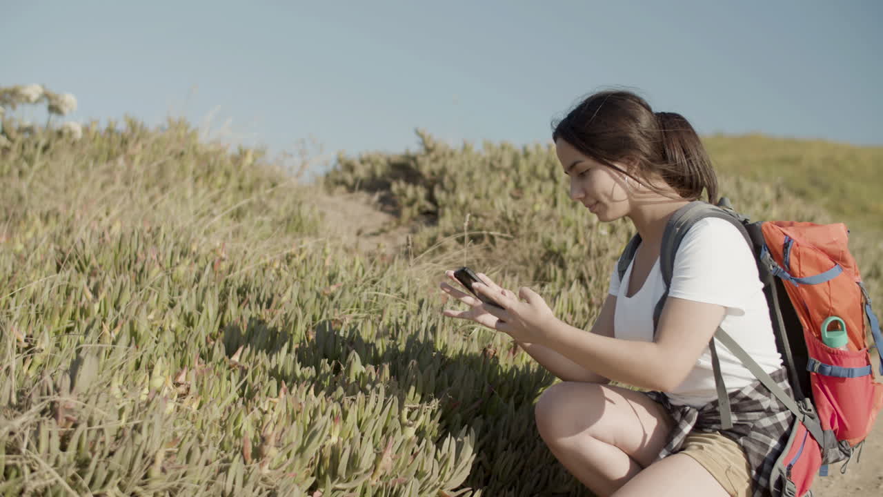 Pretty Caucasian girl with backpack taking photo of flowers with phone