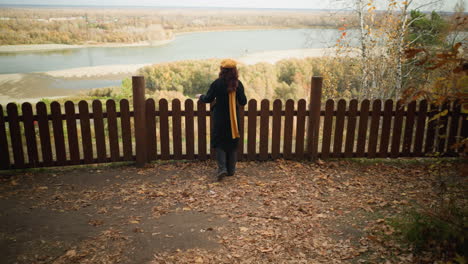 rear view of a tourist in a yellow beret and black coat walking towards a wooden fence, resting her hand on it, enjoying the scenic view of a distant river and dry autumn foliage scattered around