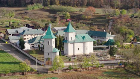 A cinematic aerial shot slowly orbits a traditional Romanian Orthodox monastery, revealing the church, green spires, a cemetery, and the surrounding autumn forest hills. Runc, Buhusi