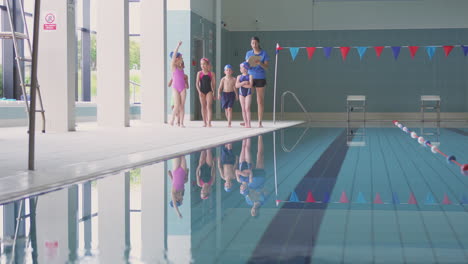 female coach walks children in swimming class along edge of indoor pool