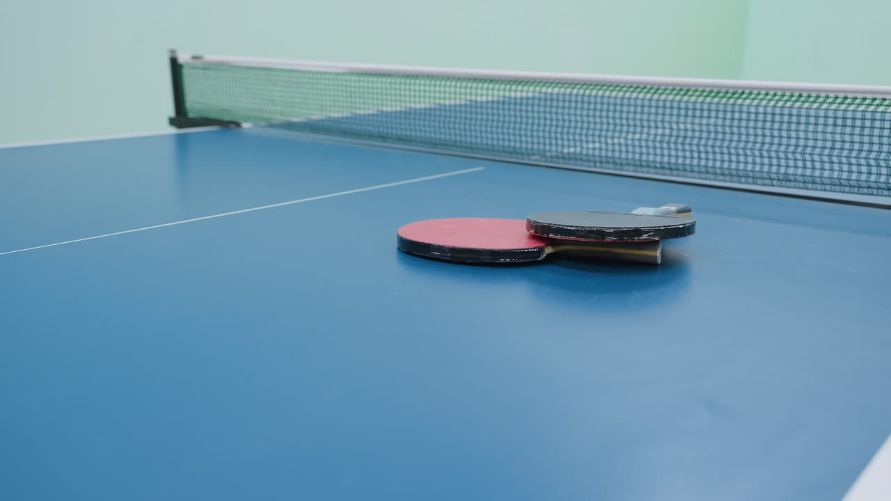 Two rackets rest on blue tennis table near net inside indoor hall ...