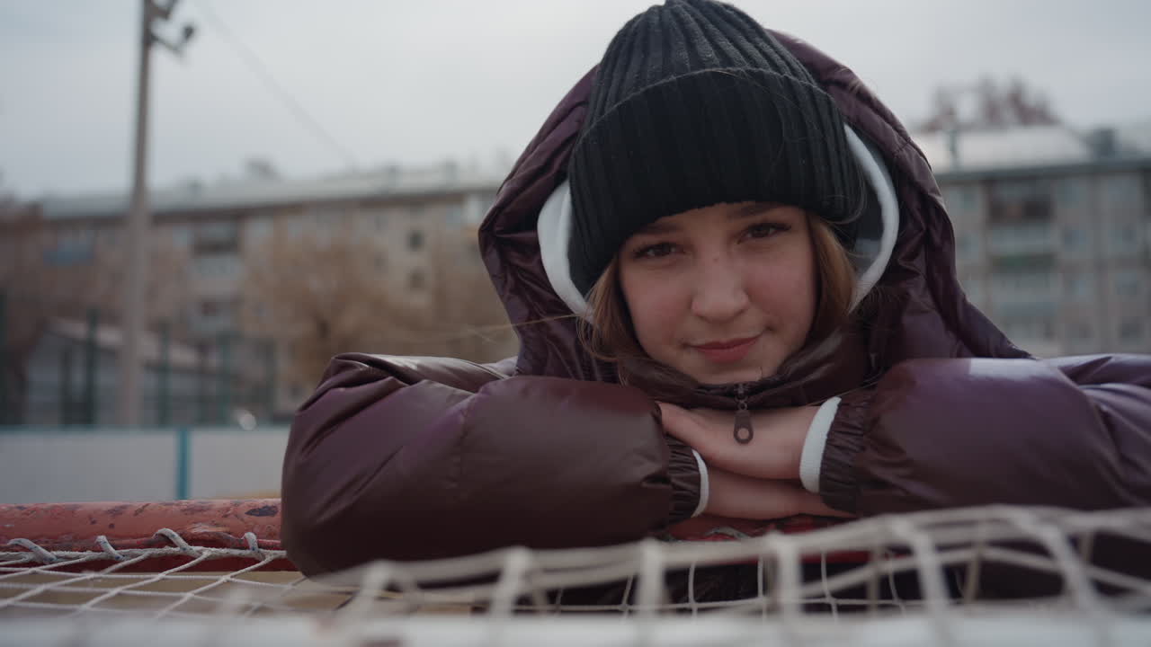 Расслабленный подросток европеоидной расы, облокотившись на сетку, смотрит нежным взглядом, в теплой куртке и шапке, позади городской корт, мягкий рассеянный свет и задумчивое настроение, тихая пауза после игры и едва заметная улыбка