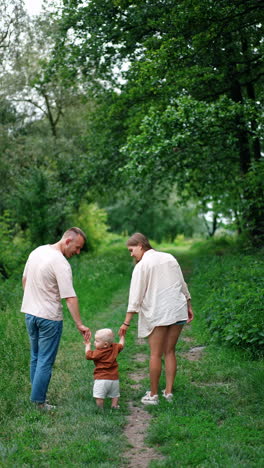mother and father leading their baby boy learning to walk. family of three in the nature in summer. vertical video.