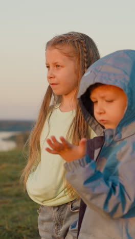 kids boy and girl at riverside in rainy evening. toddler boy looks at water drops on palm while girl stands nearby on riverbank. walking under rain