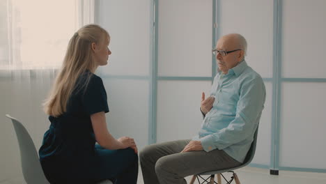 Young-Caring-Nurse-Talking-With-Very-Emotional-Elderly-Man-In-The-Office,-Holding-Hands