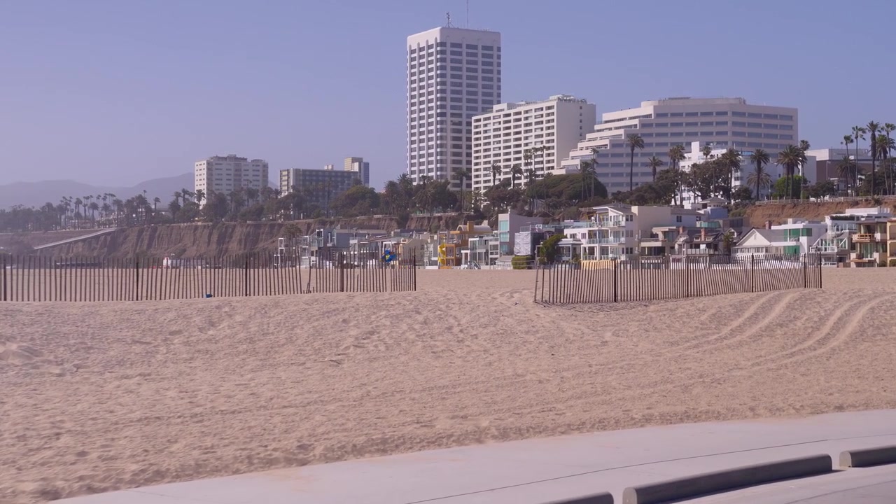 Ripresa con gimbal di una pista ciclabile sulla spiaggia a Santa Monica, CA. Mostra la spiaggia sabbiosa in primo piano e hotel famosi in lontananza.