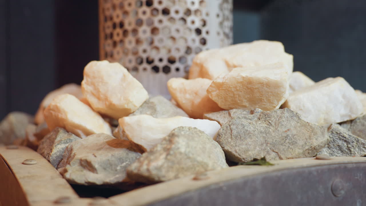 Close up of mixed sauna stones, including light and dark varieties, neatly stacked near perforated metal heater, highlighting raw textures and earthy tones in tranquil steam room environment