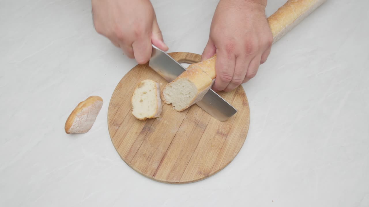 Top-down close-up of a baguette being cut on a wooden board by hand