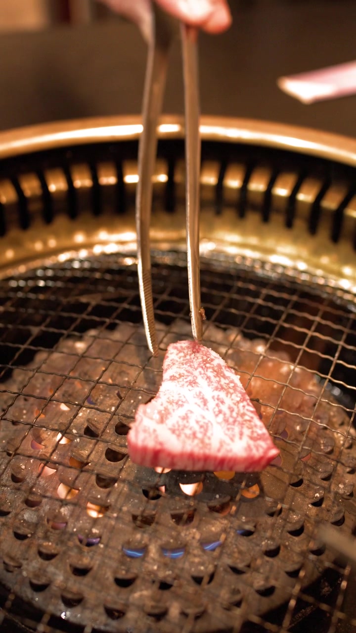 Nærbilde av marmorert biff som grilles over kullflammer i et japansk restaurantmiljø.
