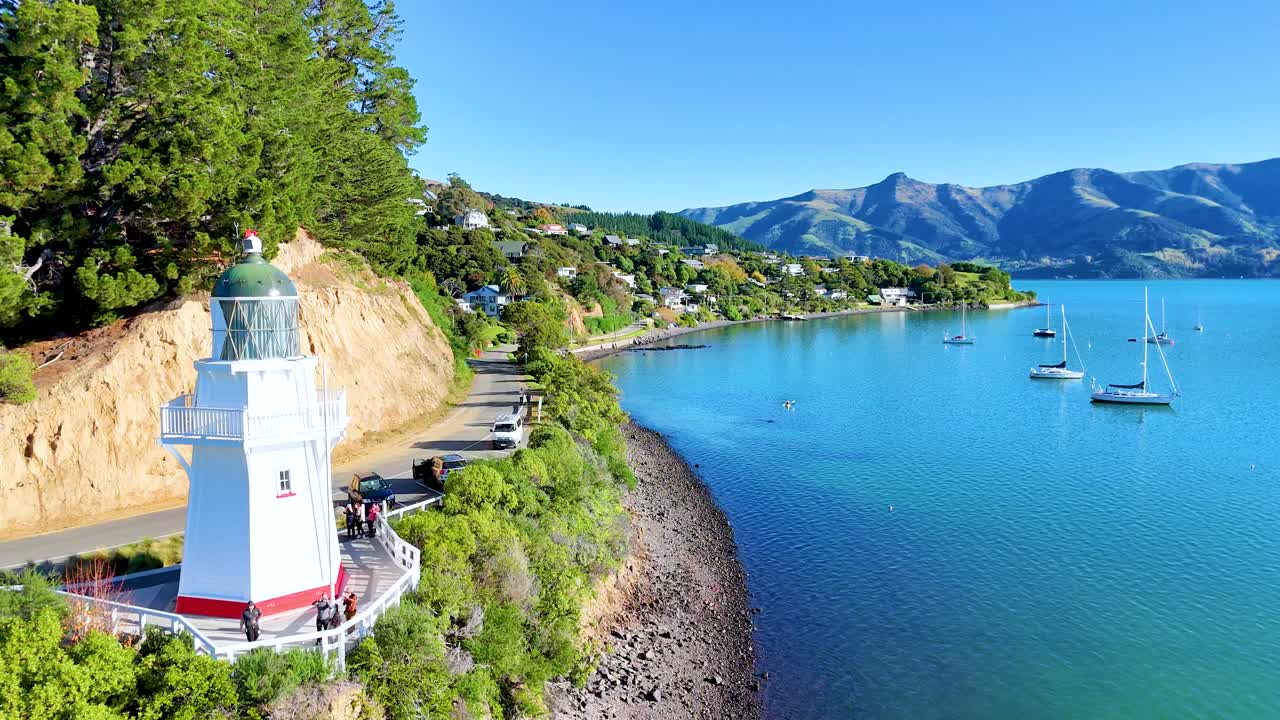 Aerial view of Akaroa's coastline with a lighthouse, clear blue waters, and sailboats under bright daylight