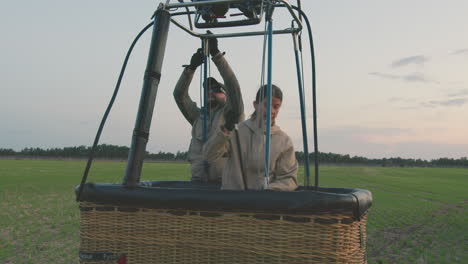 woman adjusts setup inside hot air balloon basket placed on open field with soft evening light, preparing for flight or setup activity with visible ropes and frame structure