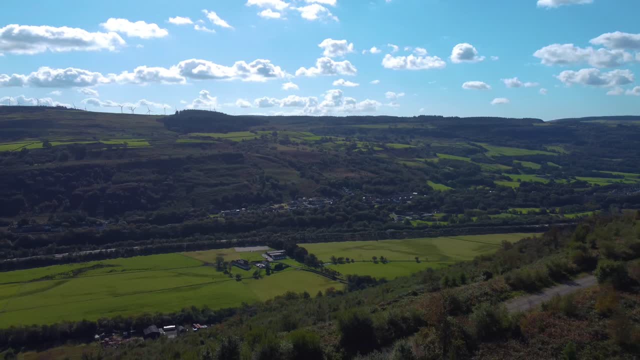 Vista aerea rotante su una valle gallese con parco eolico in cima alla collina, campi aperti e sentiero escursionistico in ghiaia