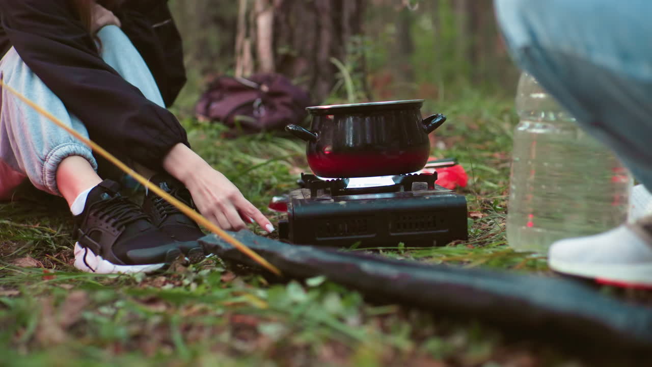 close up of two campers seated on forest ground as woman adjusts portable gas stove with black pot on top while man sits nearby on grass surrounded by gear and trees during outdoor cooking