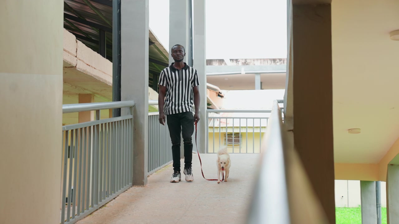 Black man walking small dog along apartment walkway in striped shirt morning sunlight leash held puppy following close calm pace metal railing residential corridor perspective candid portrait of owner