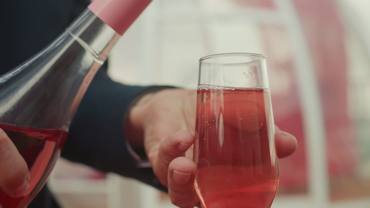 Mann, der während eines Dates auf der Terrasse Rosé in ein Glas gießt, Nahaufnahme der Flasche und der Hand, aufsteigende Bläschen, Glas fängt Licht ein, elegante Tischdeko für Paare, entspannte, festliche Stimmung