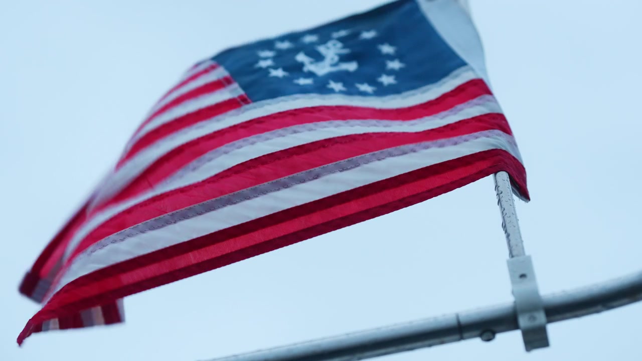 Close view of a United States yacht ensign in the wind. Red and white stripes ripple below a blue canton with white stars and an anchor above metal railings.