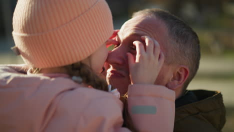 a joyful little girl in a pink cap and jacket is being carried by a man. she playfully places her hands on his eyes, creating a heartwarming and intimate moment of bonding between them