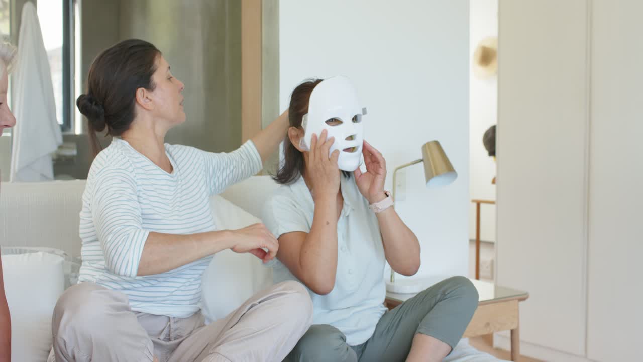 Mature daughter helping senior woman lifting white mask, brushing hair and securing fit on sofa