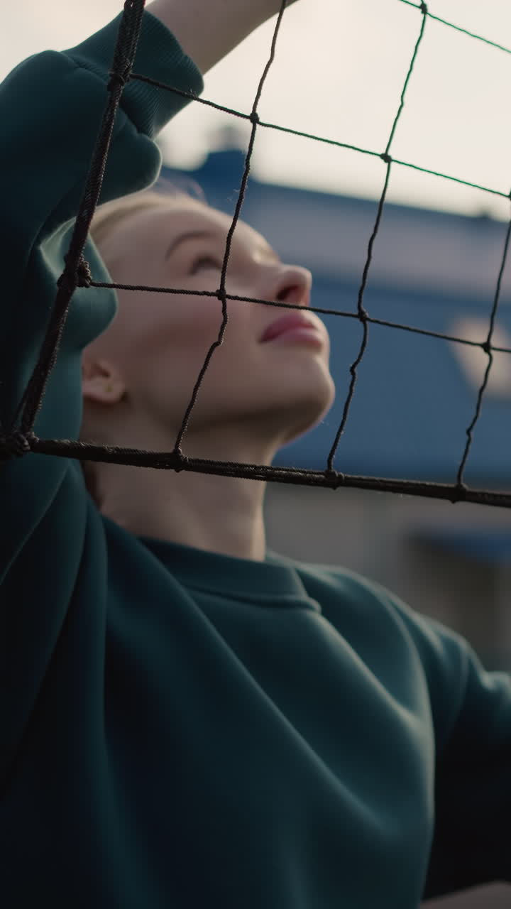 close up de una deportista con suéter verde sosteniendo y sacudiendo una red de voleibol con el fondo borroso de alguien jugando voleibol, sesión de entrenamiento deportivo al aire libre con luz solar e instalación deportiva