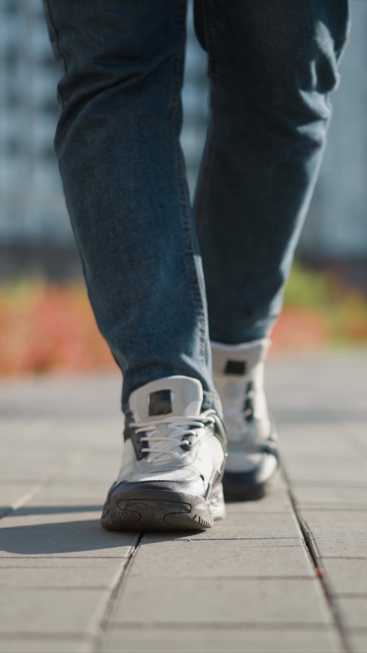 Close up of person feet in white sneakers and blue jeans walking on pavement. shoes are detailed with laces soles texture and fabric material visible as subject moves forward. Background