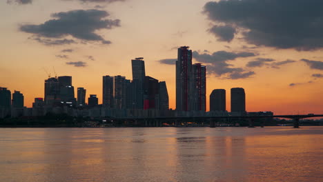 Han-River-Waterfront-and-Yeouido-Island-Skyscraper-Towers-Sihouette-Back-lit-With-Fading-Orange-Sunset-Sunlight,-Mapo-Bridge-in-Seoul-South-Korea