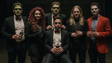 A group of creatively costumed characters, featuring classic horror aesthetics combined with formal attire, posing dramatically in a dimly lit setting while holding drinks