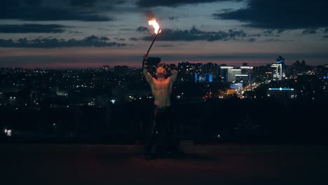 Young-blond-male-does-tricks-with-fire-breaths-fire-in-the-middle-of-the-night-with-city-skyline-in-the-background