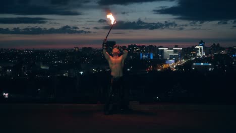 Un-Joven-Rubio-Hace-Trucos-Con-Fuego-Y-Respira-Fuego-En-Medio-De-La-Noche-Con-El-Horizonte-De-La-Ciudad-Al-Fondo