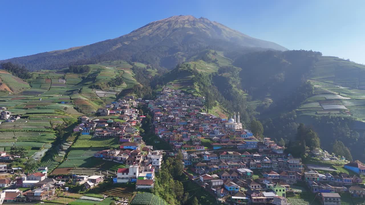 Aerial view of farming village at the slope of majestic mountain. The landscape features layered crop fields, homes, and clear blue sky, highlighting rural charm. Nepal Van Java, Indonesia