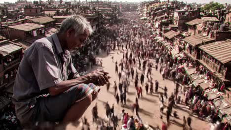 an elderly man sits quietly while people bustle around him in a crowded street of kathmandu. the atmosphere is lively with vendors selling goods amidst a rich cultural setting.