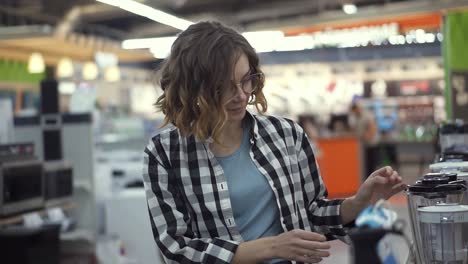 in the appliances store, a brunette curly woman in a plaid shirt chooses a blender for shopping by viewing and holding the device top in her hands. slow motion