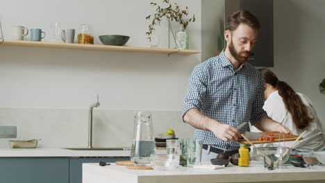 a concentrated bearded man holding chopping board while adding sliced tomatoes in glass bowl in a modern kitchen