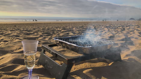 smoke sizzles from bbq grill on beach near wine glass, low angle