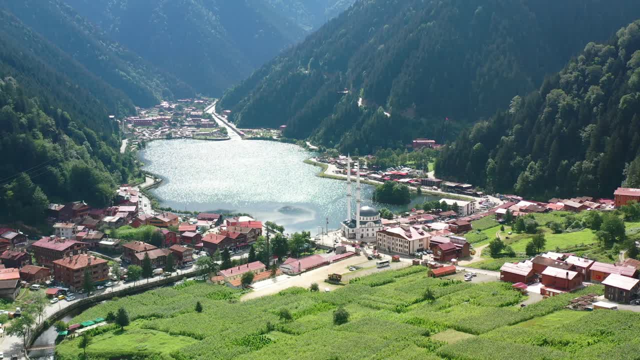 Türkiye, Trabzon, Uzungöl'de dağlarla çevrili gölden güneş yansırken aşağıdaki köyü ortaya çıkaran geriye doğru uçan drone çekimi.