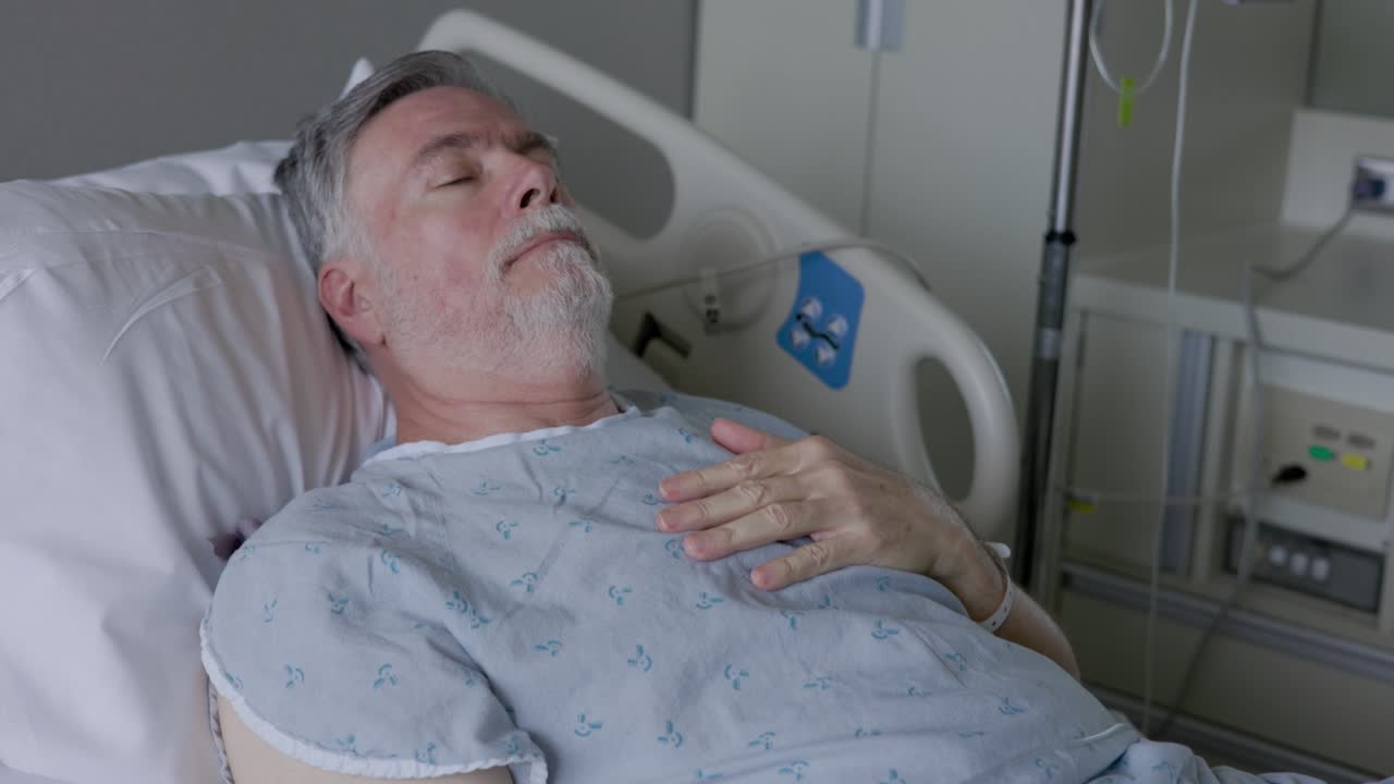 A solemn senior adult lying in a hospital bed hooked up to an IV solution delivered via a catheter or a needle in the his vein.