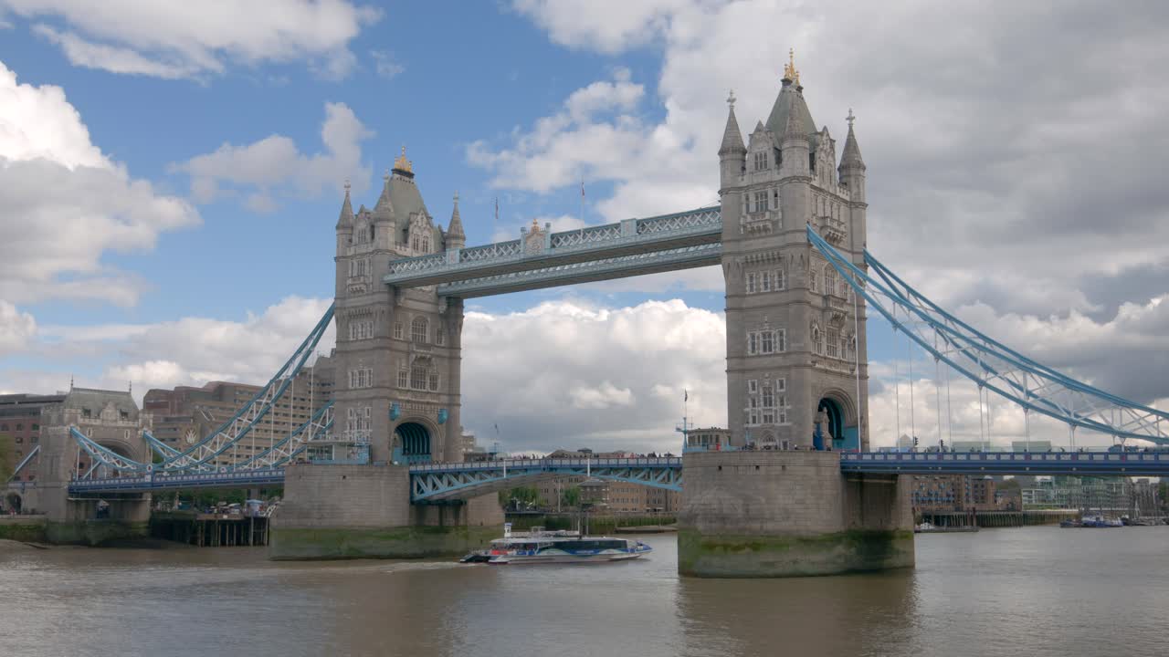 La técnica de timelapse captura el Tower Bridge extendiéndose sobre el río Támesis en Londres, con torres gemelas góticas, elementos de suspensión azules y reflejos en el agua que fluye debajo.