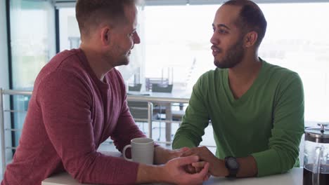 multi ethnic gay male couple talking holding hands in kitchen