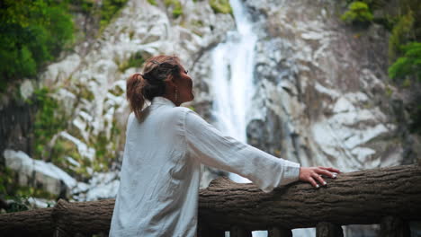 Woman-with-dark-blonde-hair-stands-in-front-of-a-waterfall,-leaning-on-a-wooden-railing