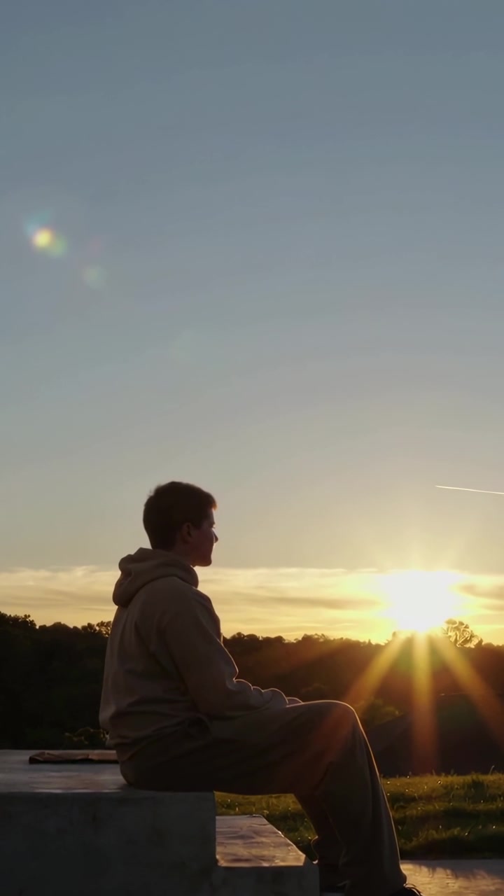 Video verticale: Adolescente seduto con felpa con cappuccio che guarda il sole tramontare, riflettendo in un punto panoramico, con il telefono accanto.
