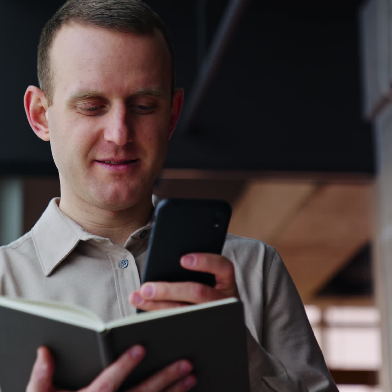 Mature man holds the notebook opened in front of him and print something on the phone. He throws interested look into the window