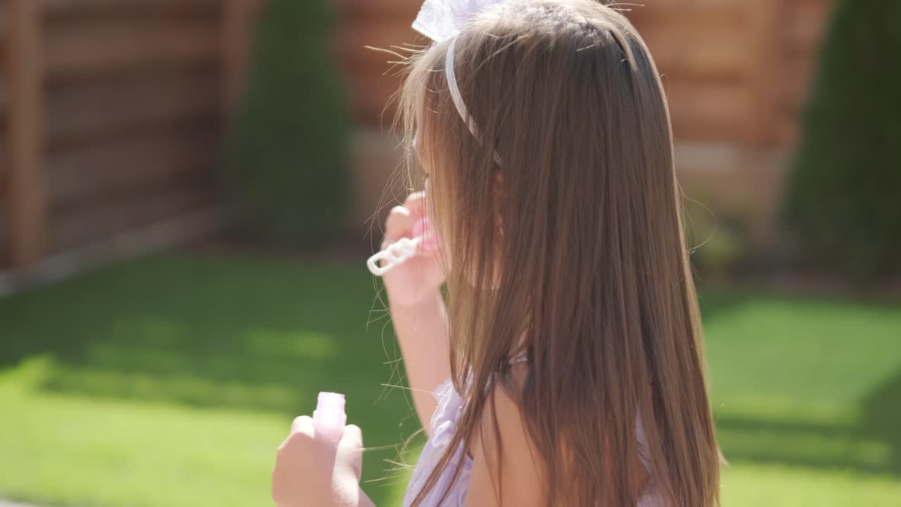 A little girl in a dress is blowing soap bubbles in the backyard on a sunny summer day
