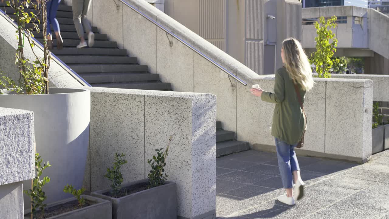 Donna adulta con giacca verde che cammina su una terrazza verso le scale, fermandosi per rispondere a una telefonata con un caffè in mano.