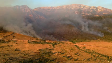 Aerial-footage-of-dramatic-wildfires-burning-in-the-mountains-on-the-outskirts-of-the-city-of-Vlorë,-Albania,-lit-by-an-orange-sunset